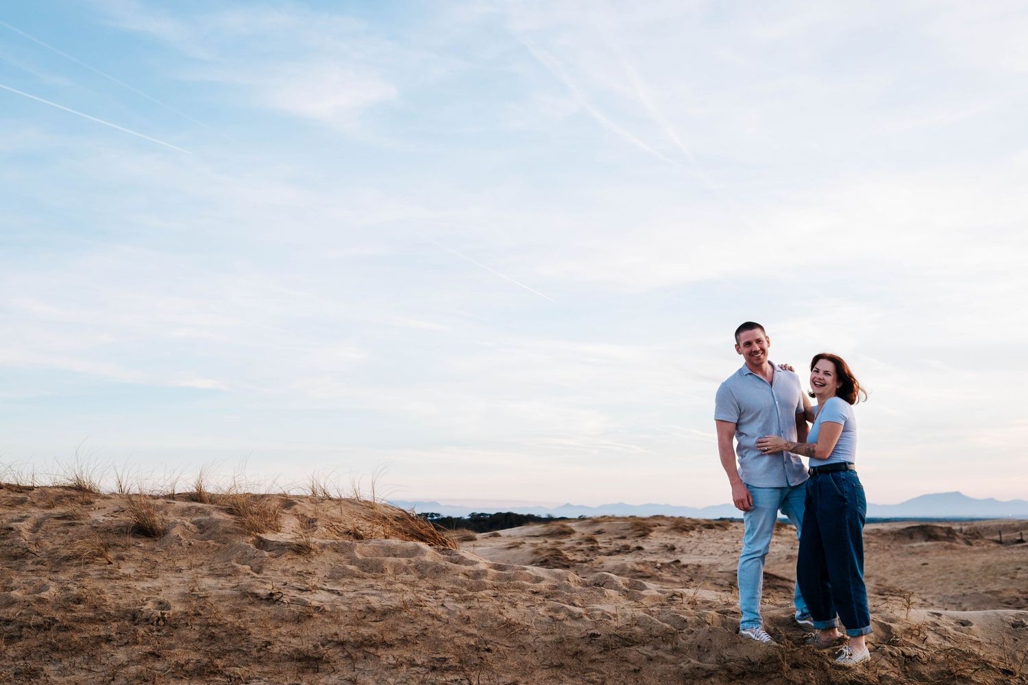 A couple stands on a rocky desert cliff during sunset overlooking a vast desert landscape.