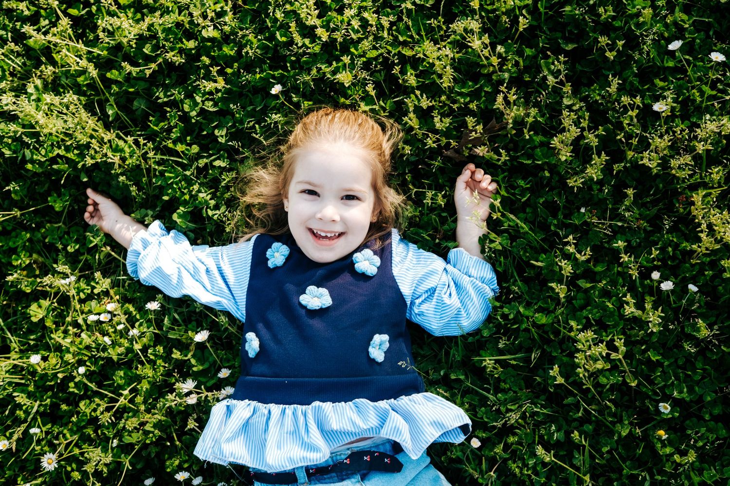 Young girl in blue floral dress smiling while lying on lush green grass with daisies on a sunny day.