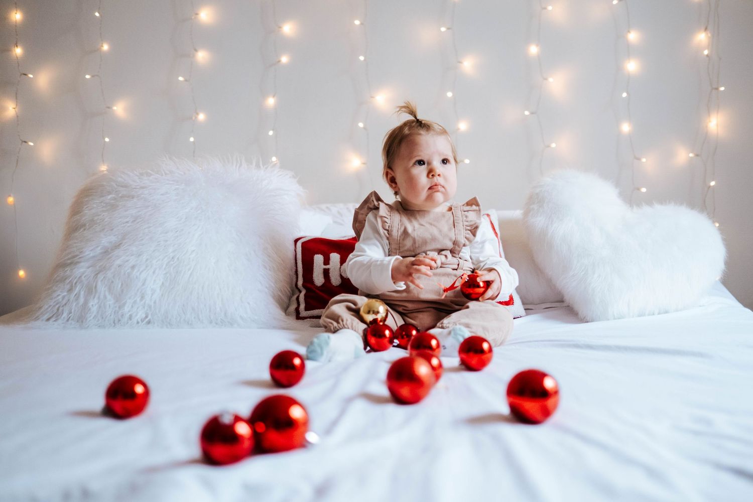 A baby lies on white bedding surrounded by red Christmas ornaments and twinkling holiday lights in the background.