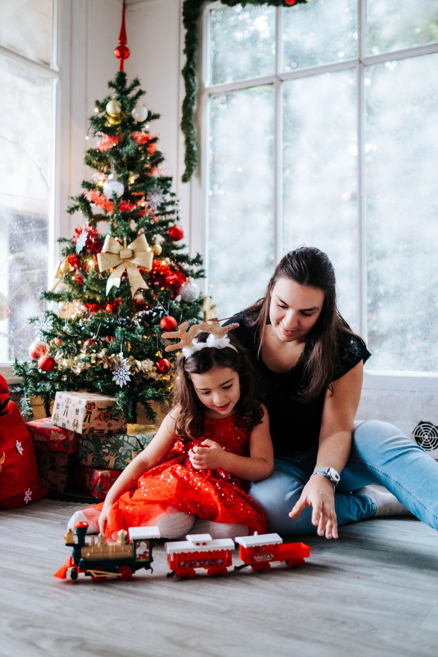 People laying on floor near decorated Christmas trees with presents and holiday decorations in bright sunlit room.