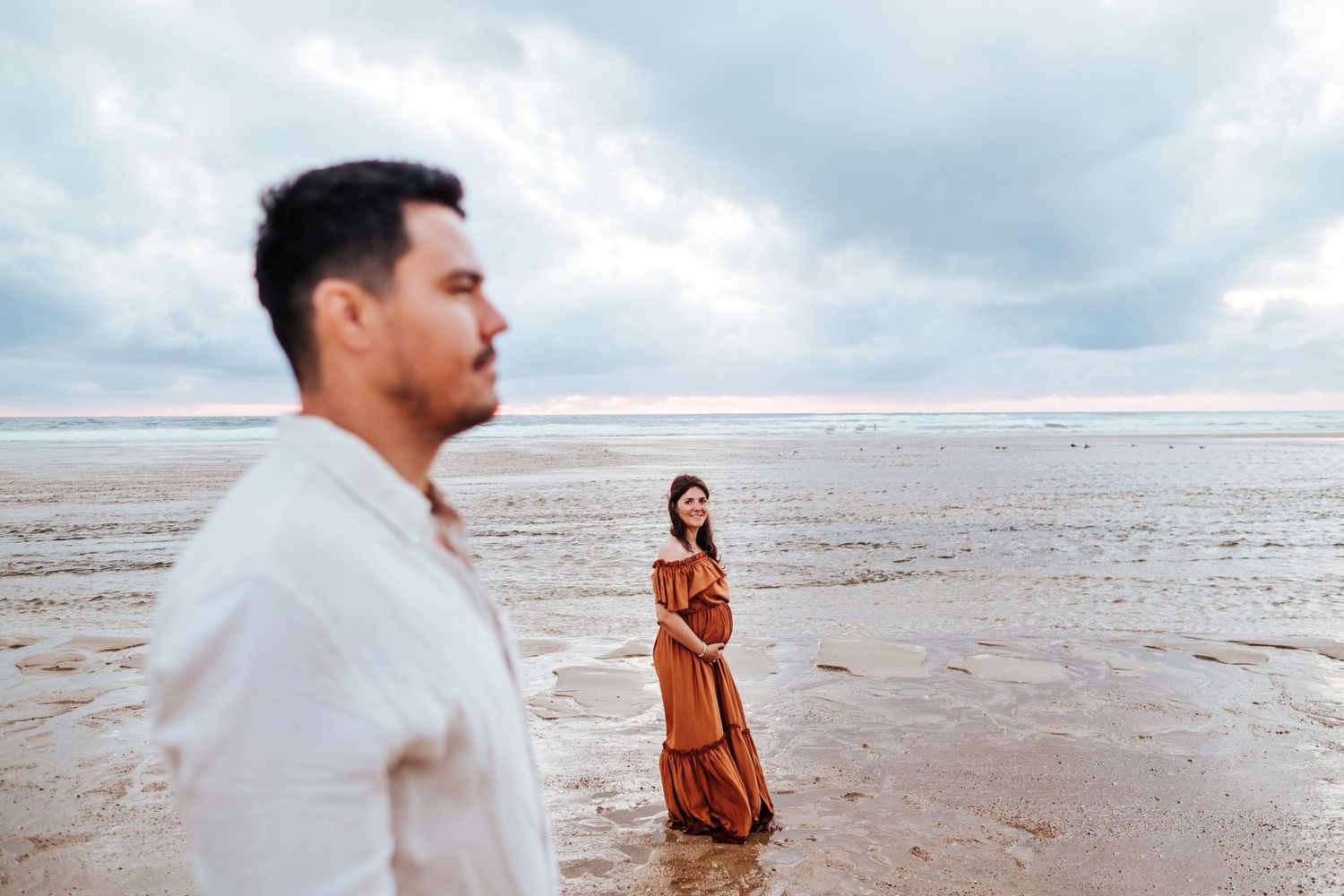 A person in white clothing stands in shallow ocean water while another figure in orange appears in the distance.