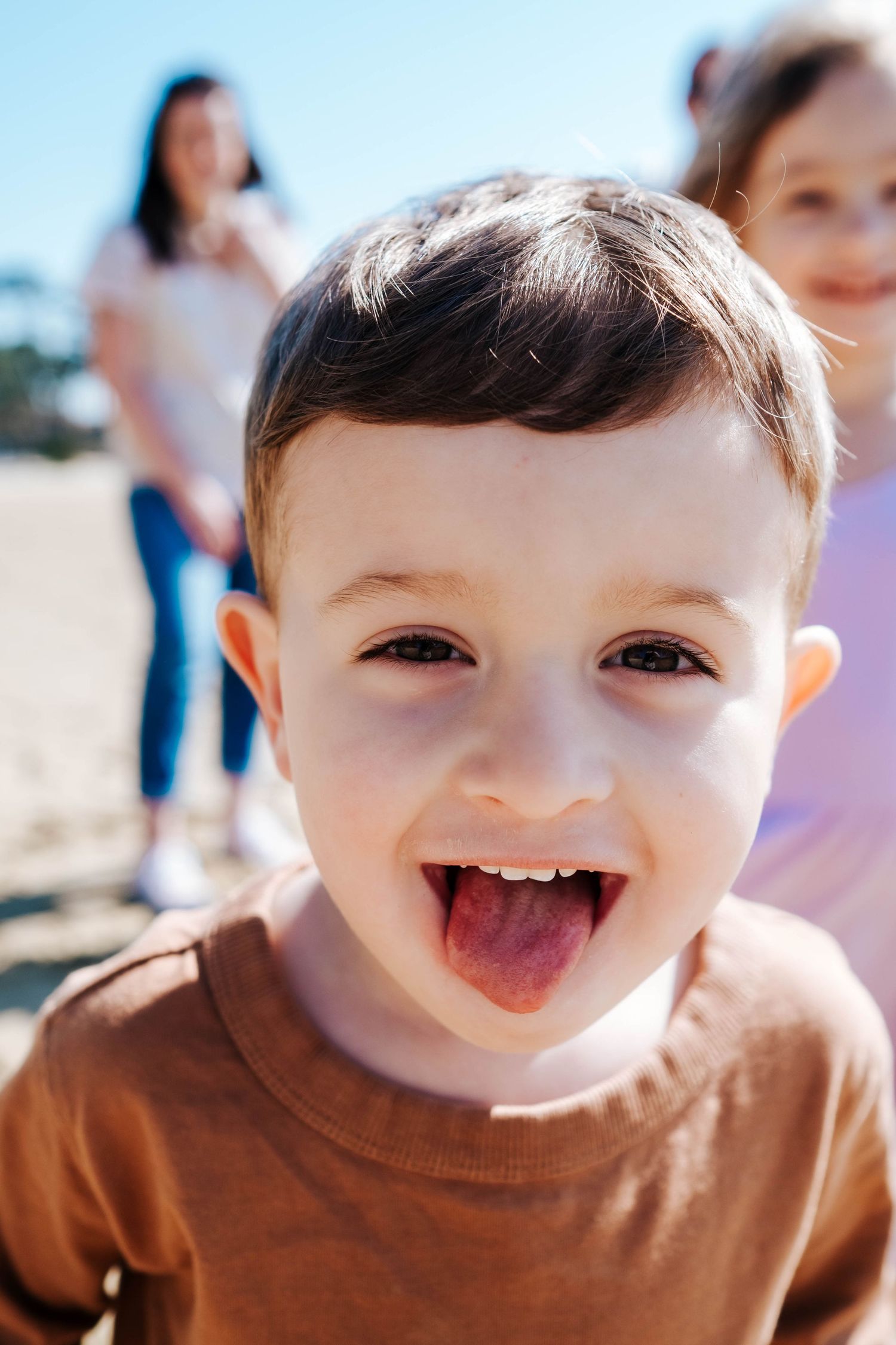 A happy toddler boy in a brown shirt sticks out his tongue at the beach with family in the background.
