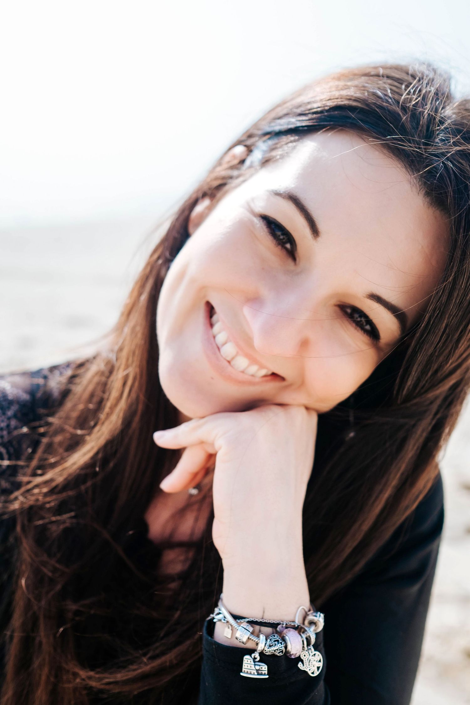 Smiling young woman with long brown hair resting chin on hand, wearing a charm bracelet, outdoors in bright sunlight.