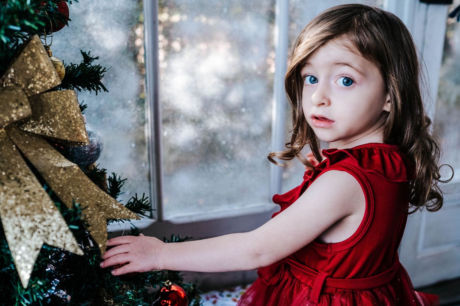 Child in red velvet dress decorates a Christmas tree with gold ornaments and twinkling lights.
