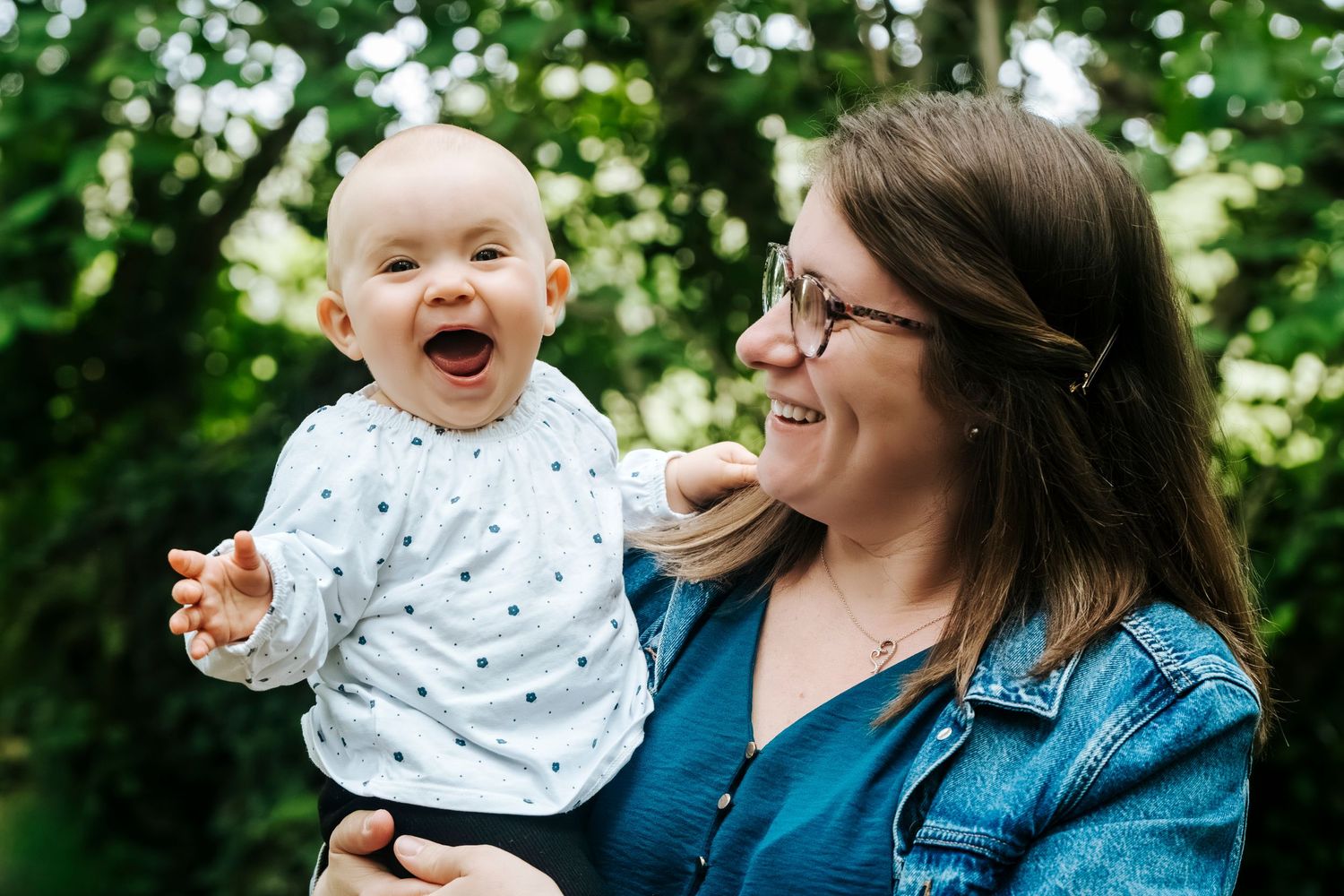 A joyful baby laughing while being held by a smiling woman with glasses outdoors among green trees.
