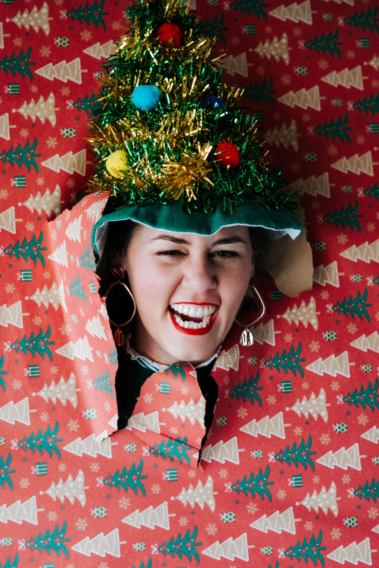 Someone peeks through Christmas wrapping paper wearing a festive tinsel crown against a red background with tree patterns.