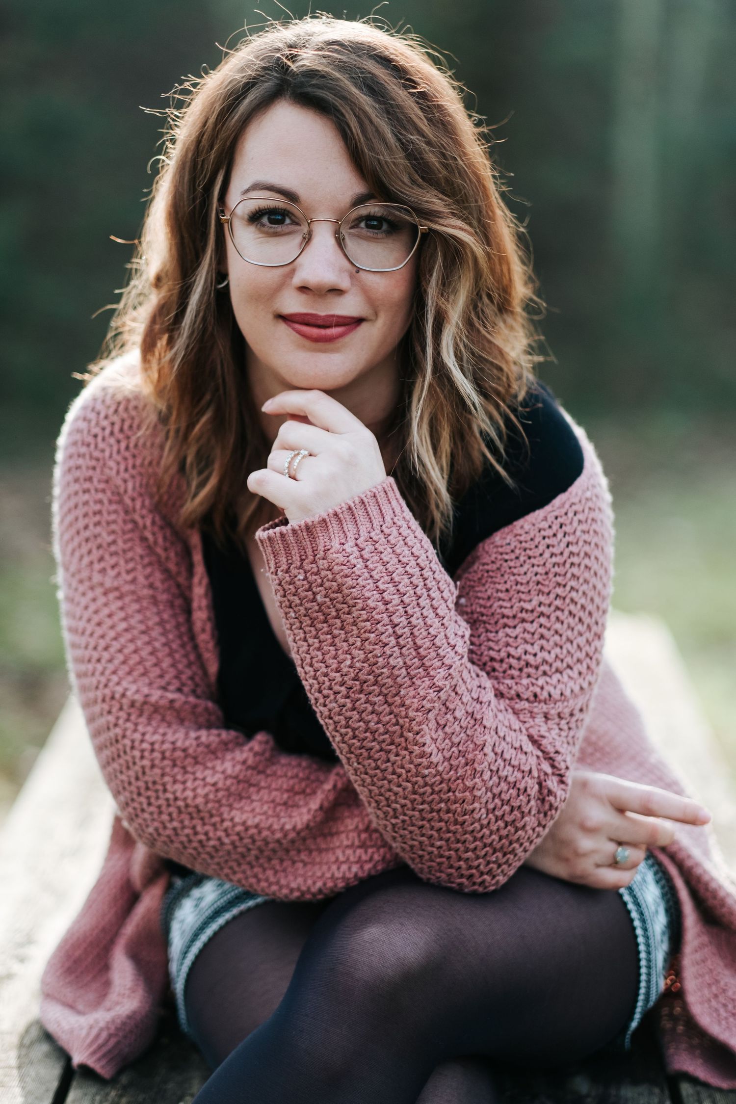 Portrait of someone in a pink knit sweater and glasses sitting outdoors with a thoughtful expression.