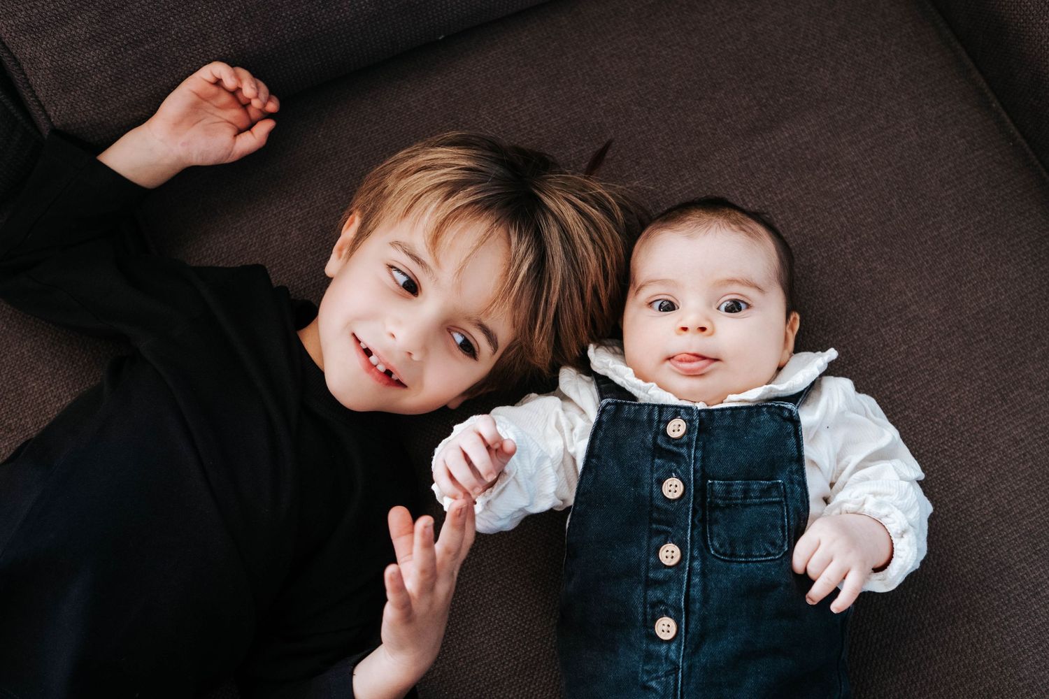 Two children, a toddler boy and baby girl, lying on a brown sofa, smiling at the camera.
