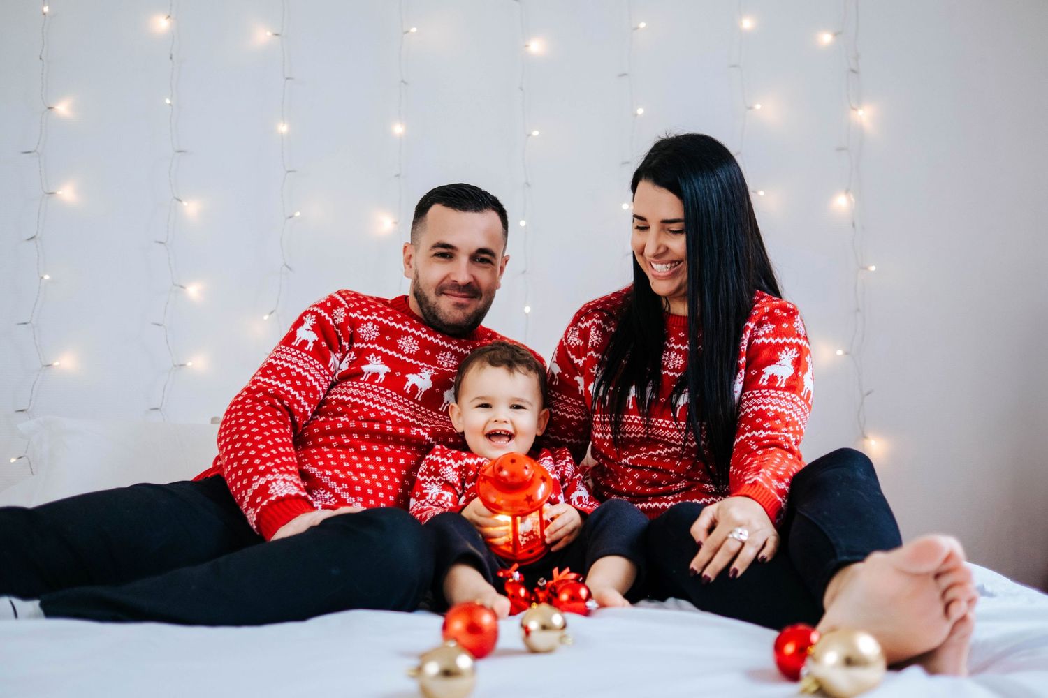 A family wearing matching red Christmas sweaters sits together on white bedding with twinkle lights in background.
