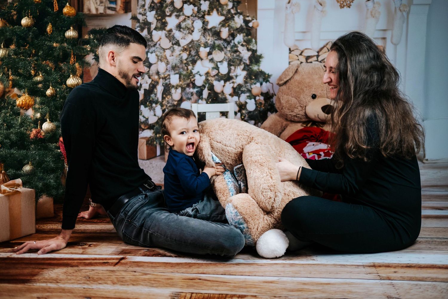 Family sits on floor by Christmas tree with large teddy bear during holiday celebration at home.