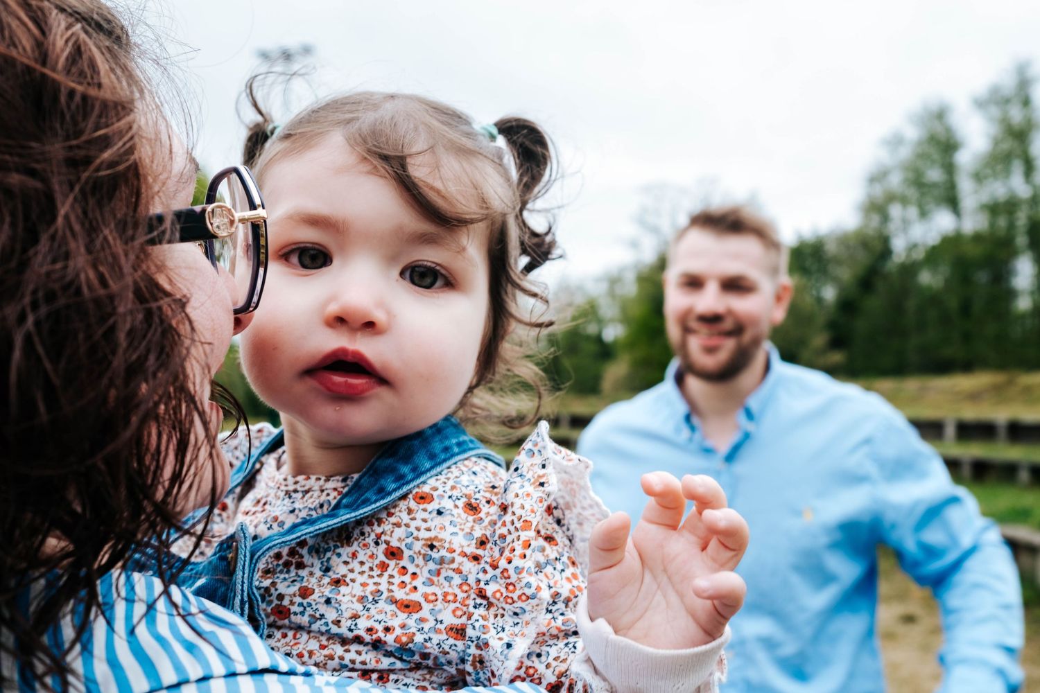 A smiling family outdoors, with a mother holding a curious baby girl with pigtails while the father smiles in the background.