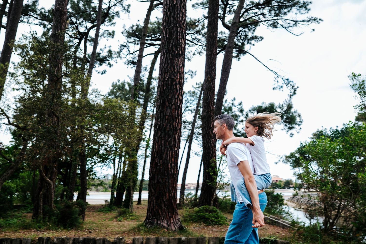 Couple walking through a pine forest near a lake, man carrying woman on his back, sharing a joyful outdoor moment.