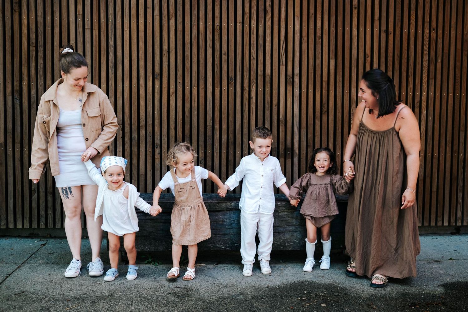 Two women hold hands with four young children in front of a wooden wall backdrop.