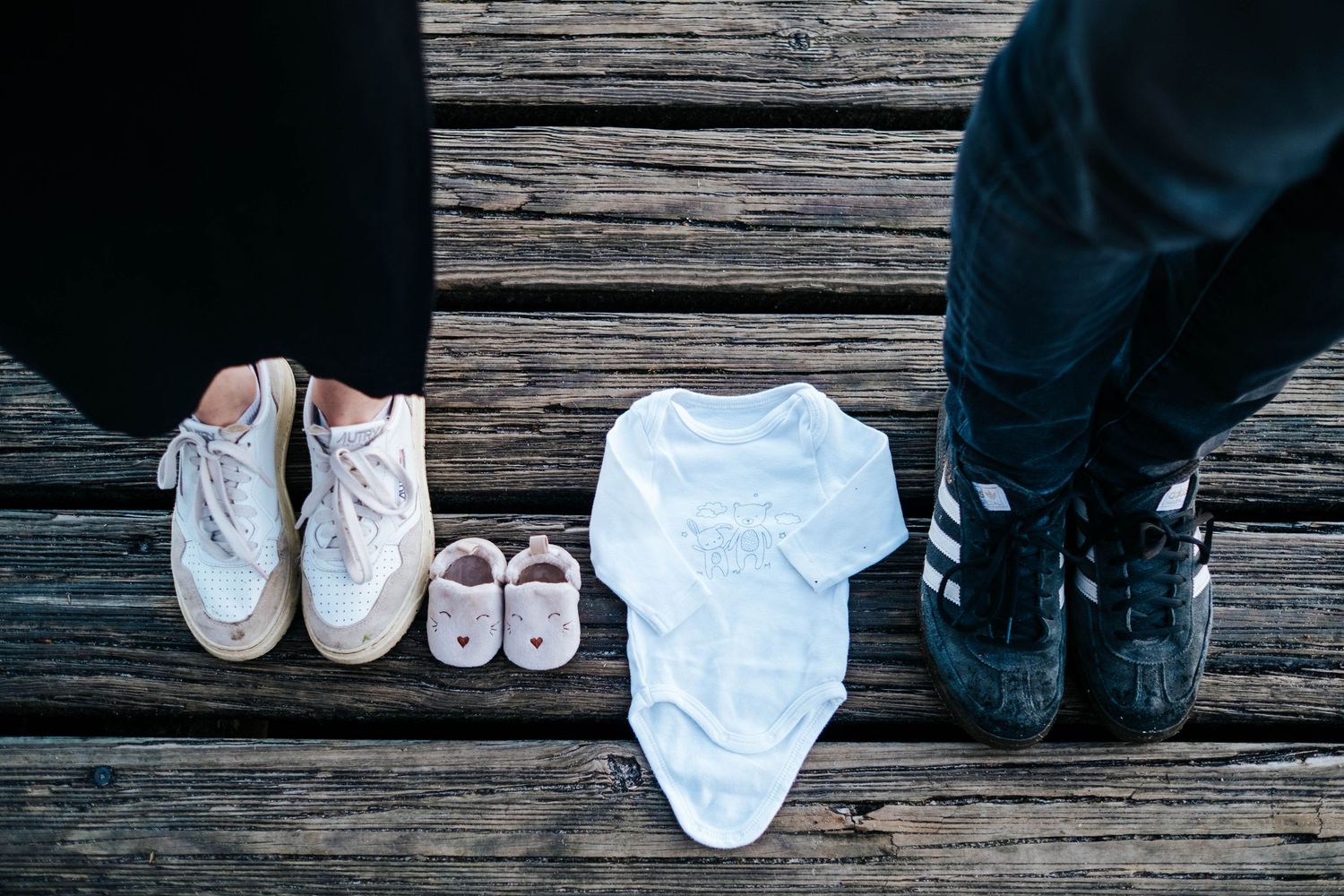 Parents stand beside tiny baby shoes and white onesie on wooden deck announcing pregnancy.