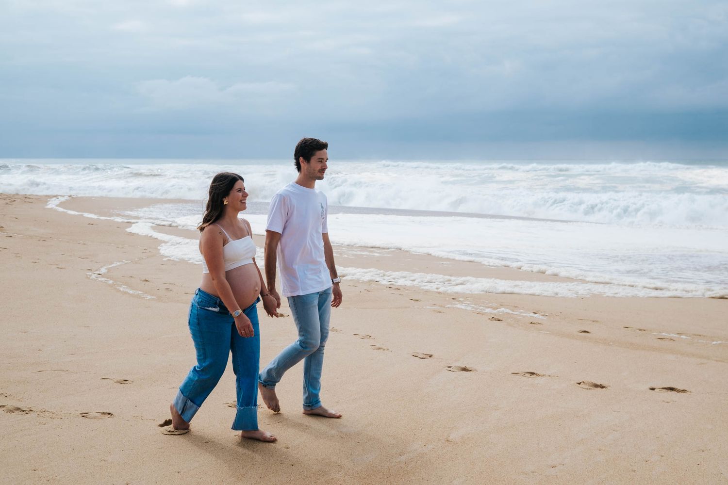 A couple walks together along a serene beach shoreline with ocean waves and cloudy skies in the background.