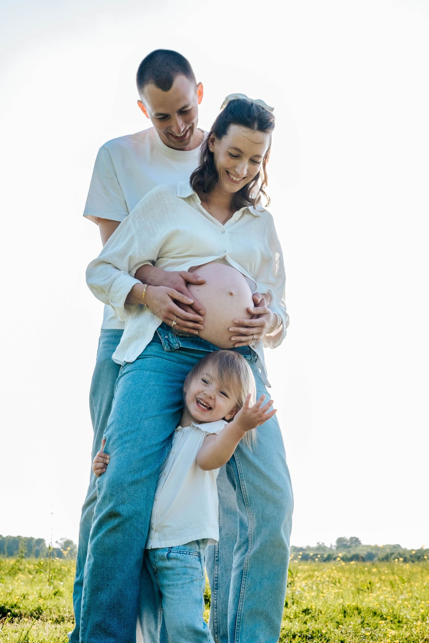 Happy pregnant woman with husband and young child embracing outdoors in a sunny green field.