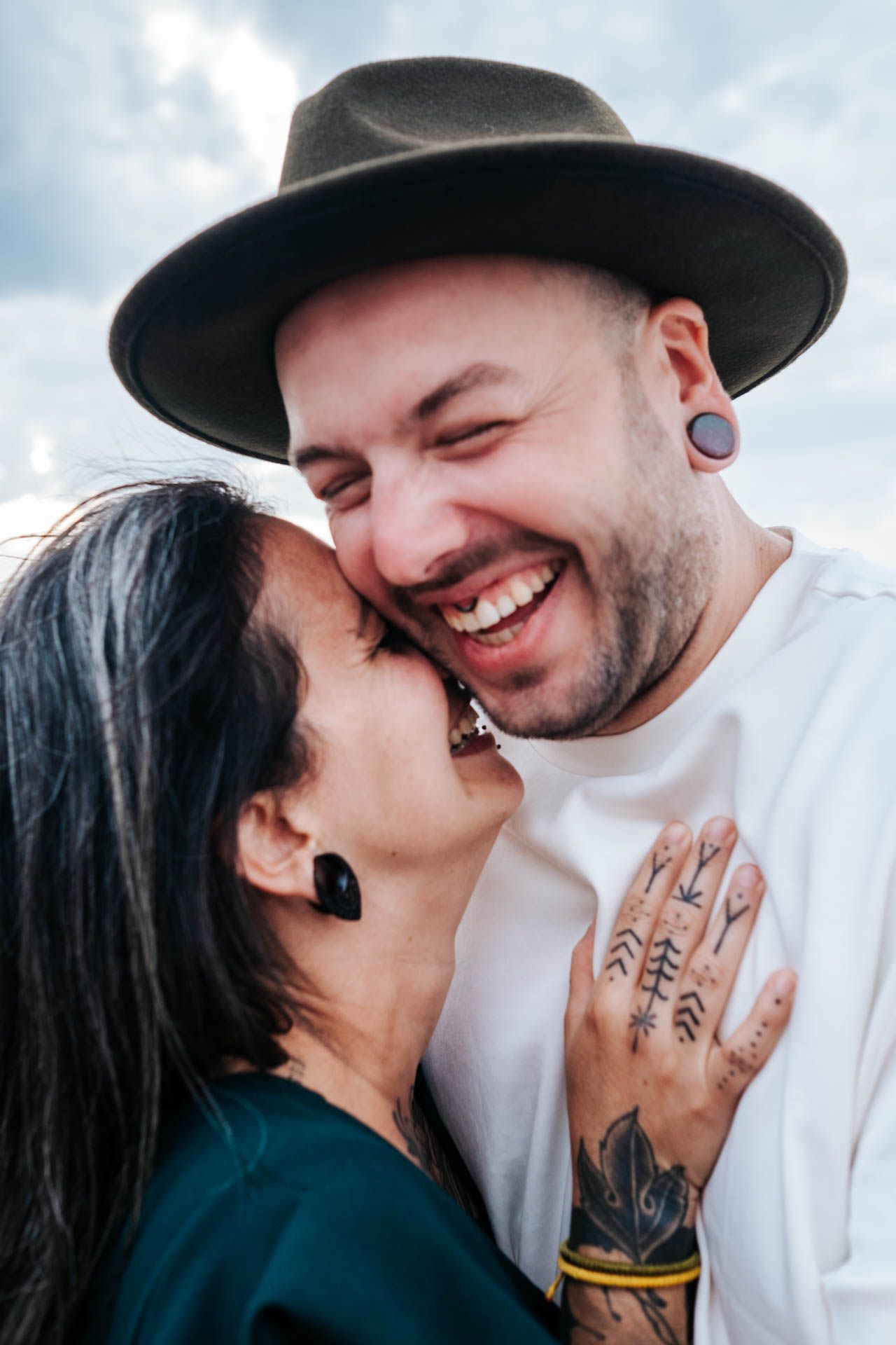 Tattooed couple sharing a joyful moment, man in wide-brim hat with gauge earrings laughing as woman kisses his cheek.