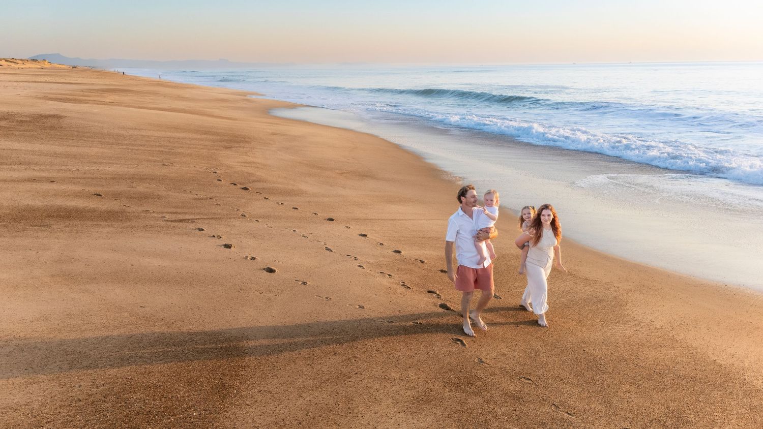Family walking on sandy beach at sunset, waves in background, wearing white outfits, footprints in sand.