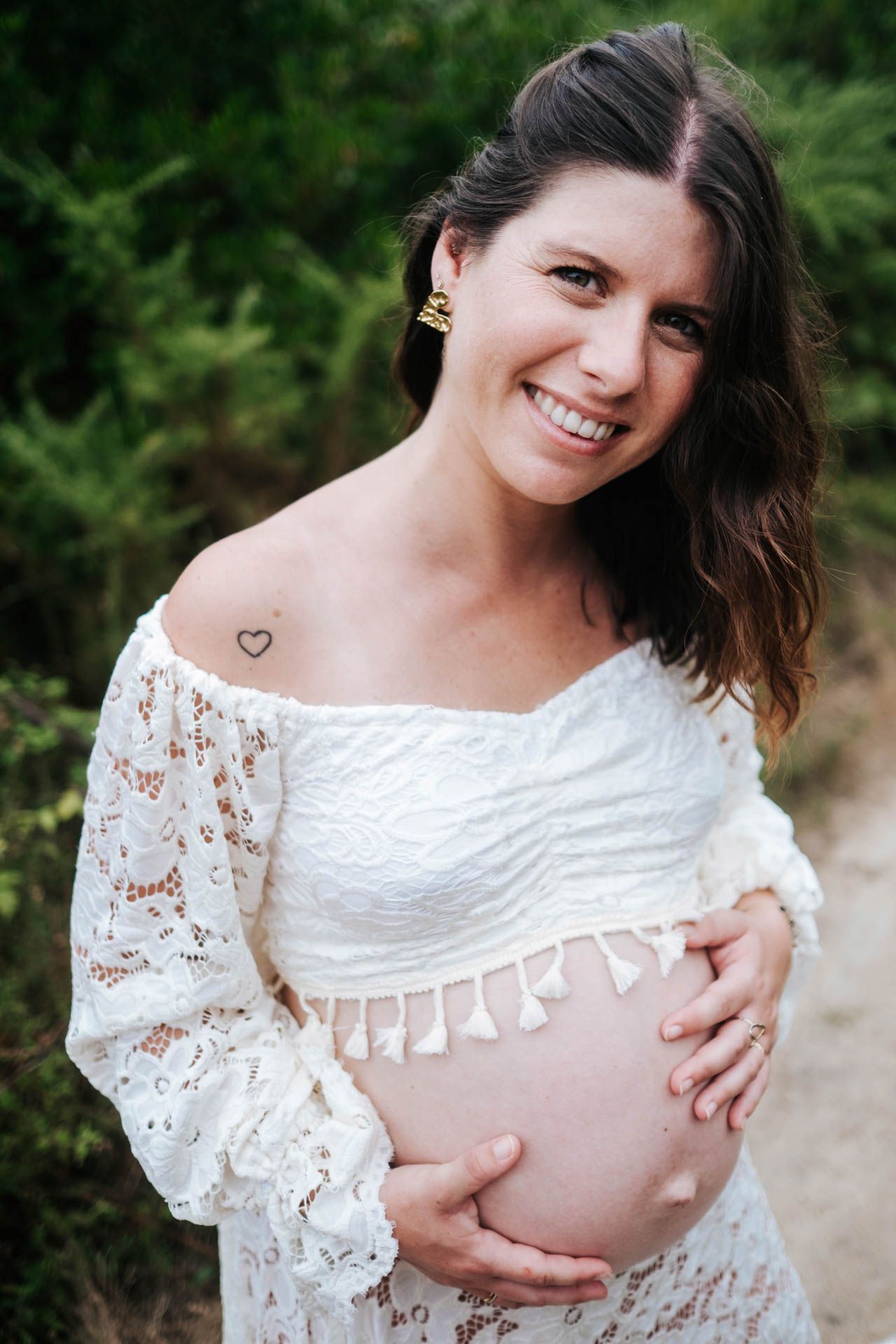 Maternity photos in white lace dress showing pregnant belly in outdoor natural setting.