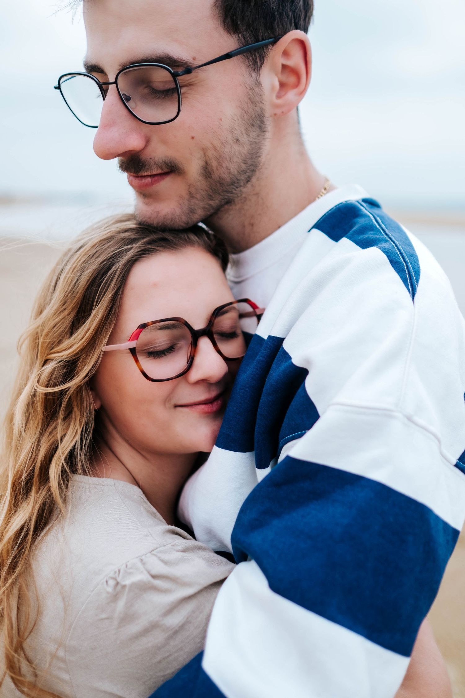 A couple wearing matching glasses embrace tenderly at the beach, dressed in white and blue striped clothing.