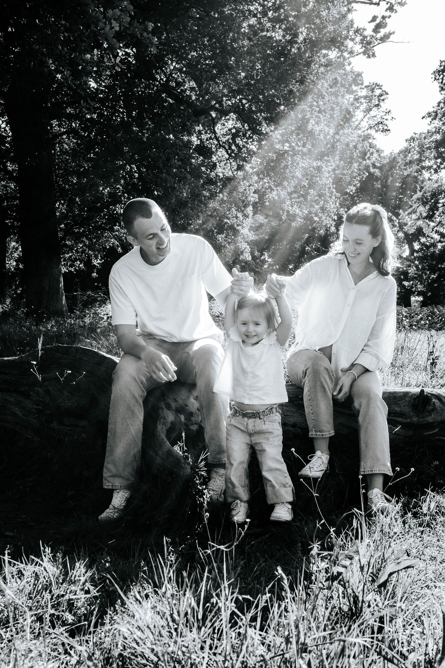 Black and white photo of a happy family with a toddler sitting on a log outdoors surrounded by trees.