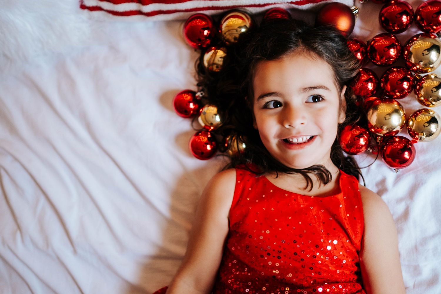 A festive holiday scene with someone lying on white fabric surrounded by red and gold Christmas ornaments and decorations.
