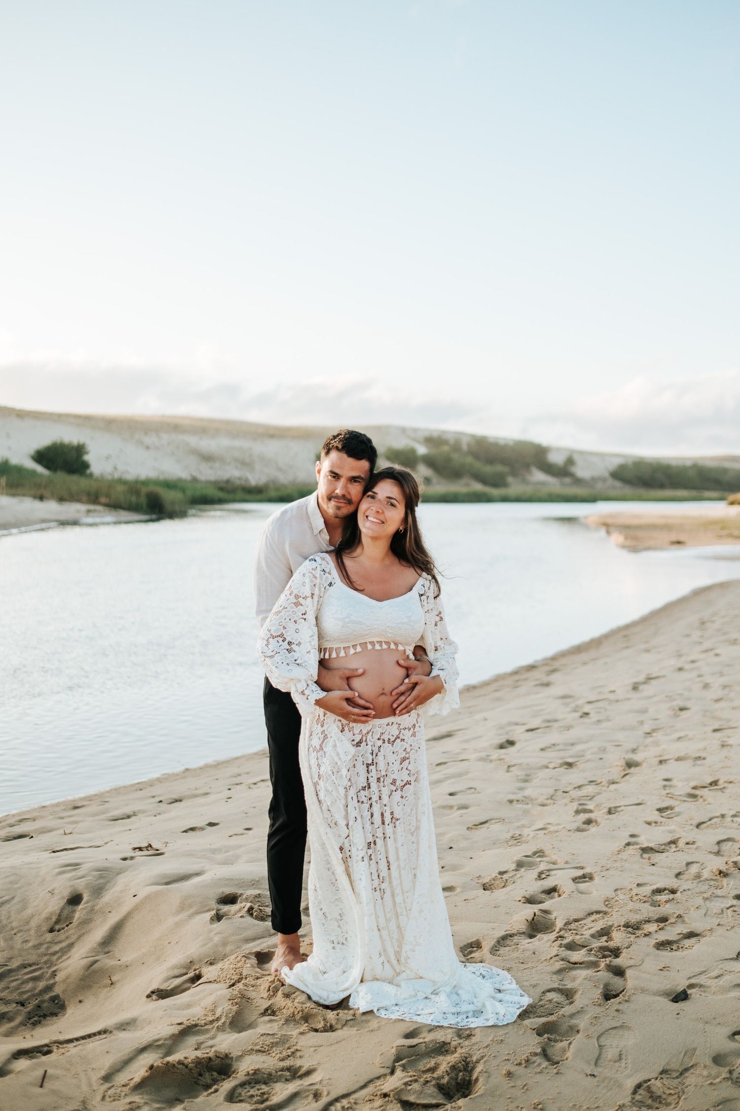 A couple in white lace stands together on a sandy beach at sunset with water and rolling hills in the background.