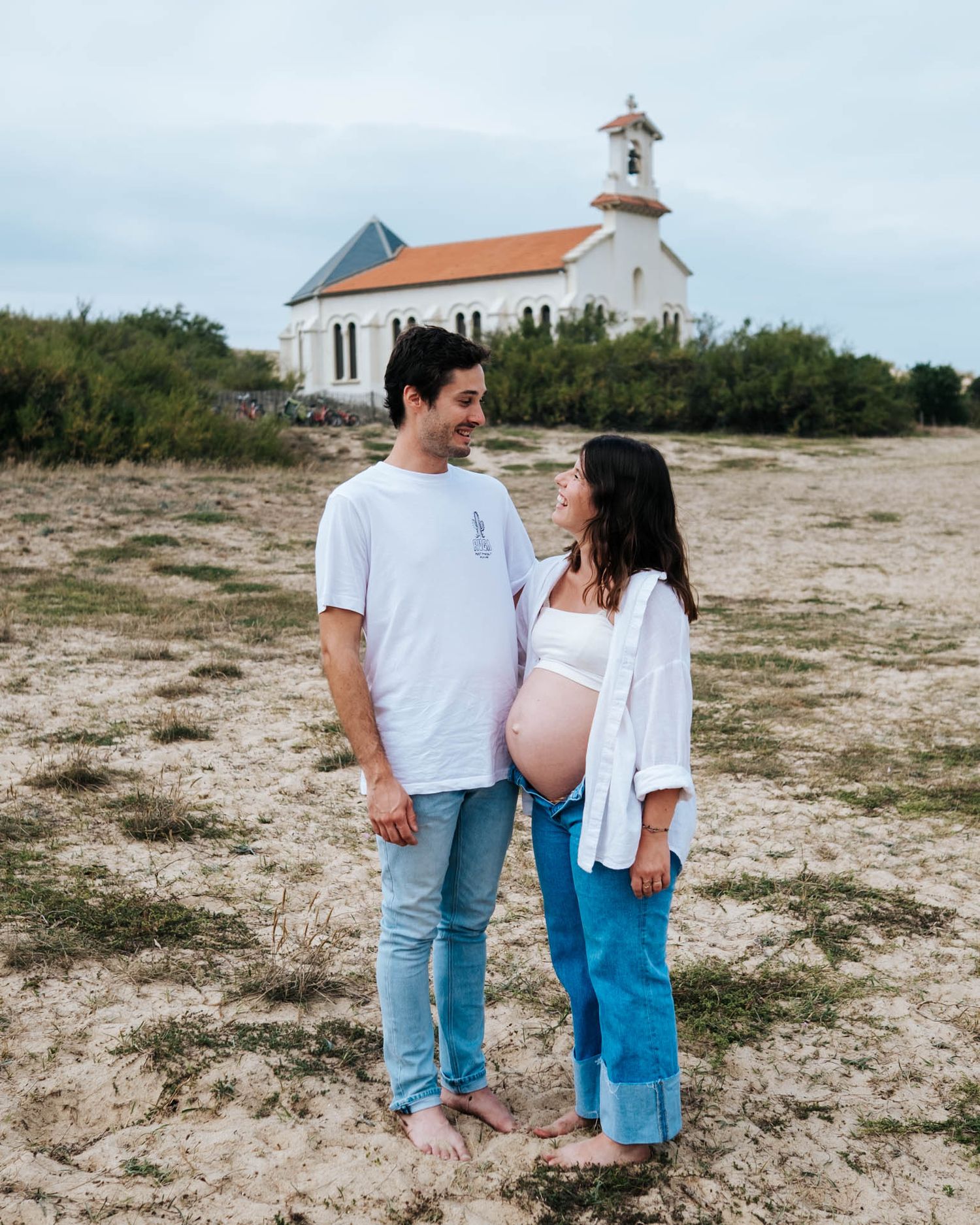 A couple in white shirts and jeans walking together on a sandy beach with a lighthouse in the background.