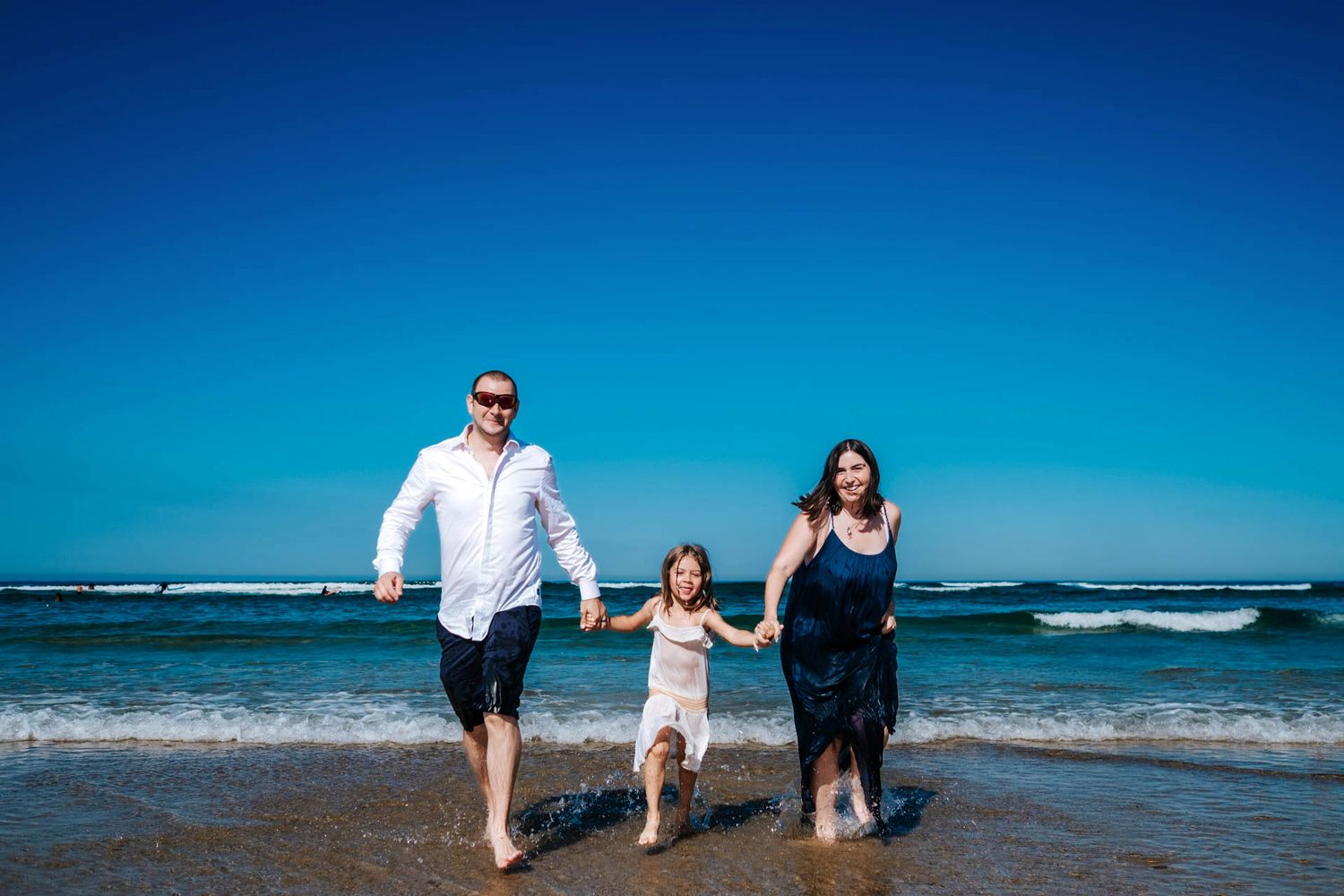Happy family of three running barefoot on a sunny beach with blue sky and ocean waves.