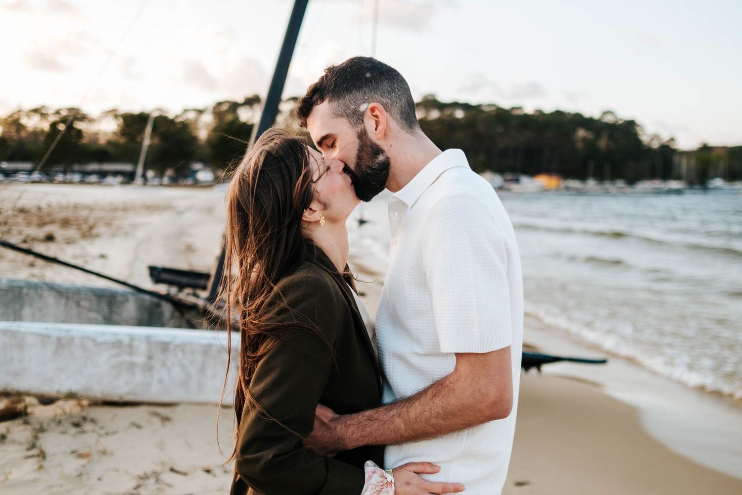 Couple sharing a romantic kiss on a sandy beach near the water at sunset, with trees in the background.