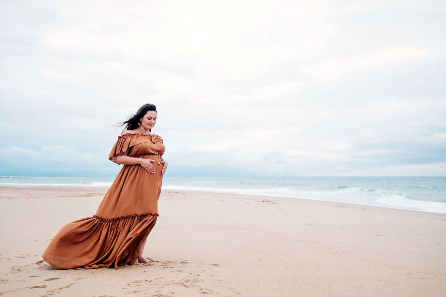 A person in a flowing rust-colored dress sits gracefully on a sandy beach with ocean waves in the background.