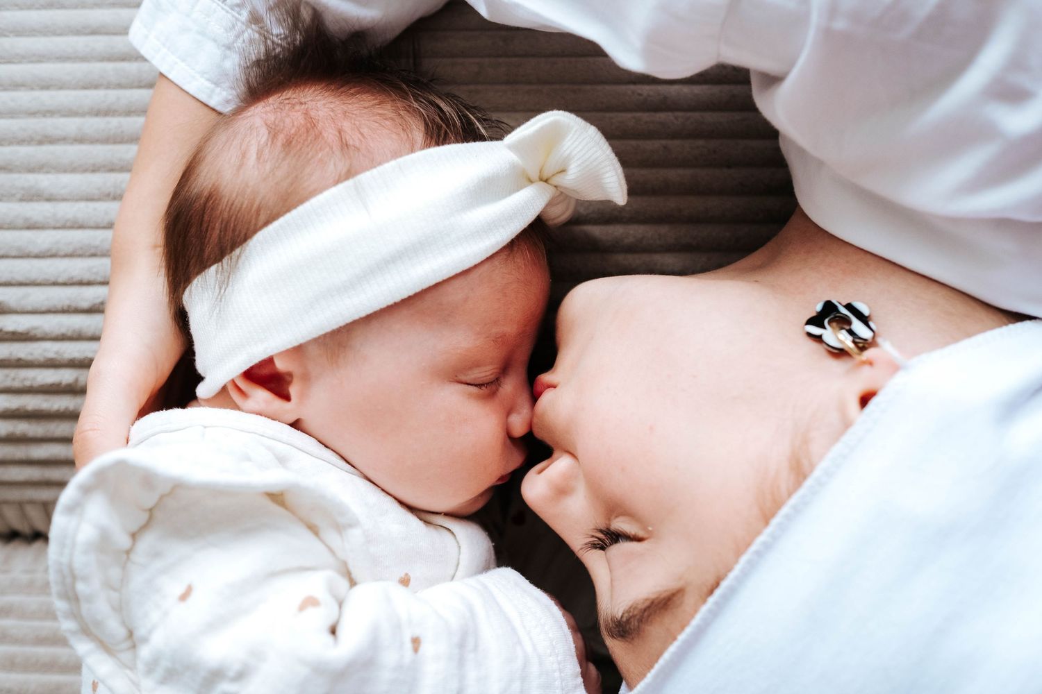 A newborn baby in a white headband snuggles close to a smiling mother while lying together on a soft surface.
