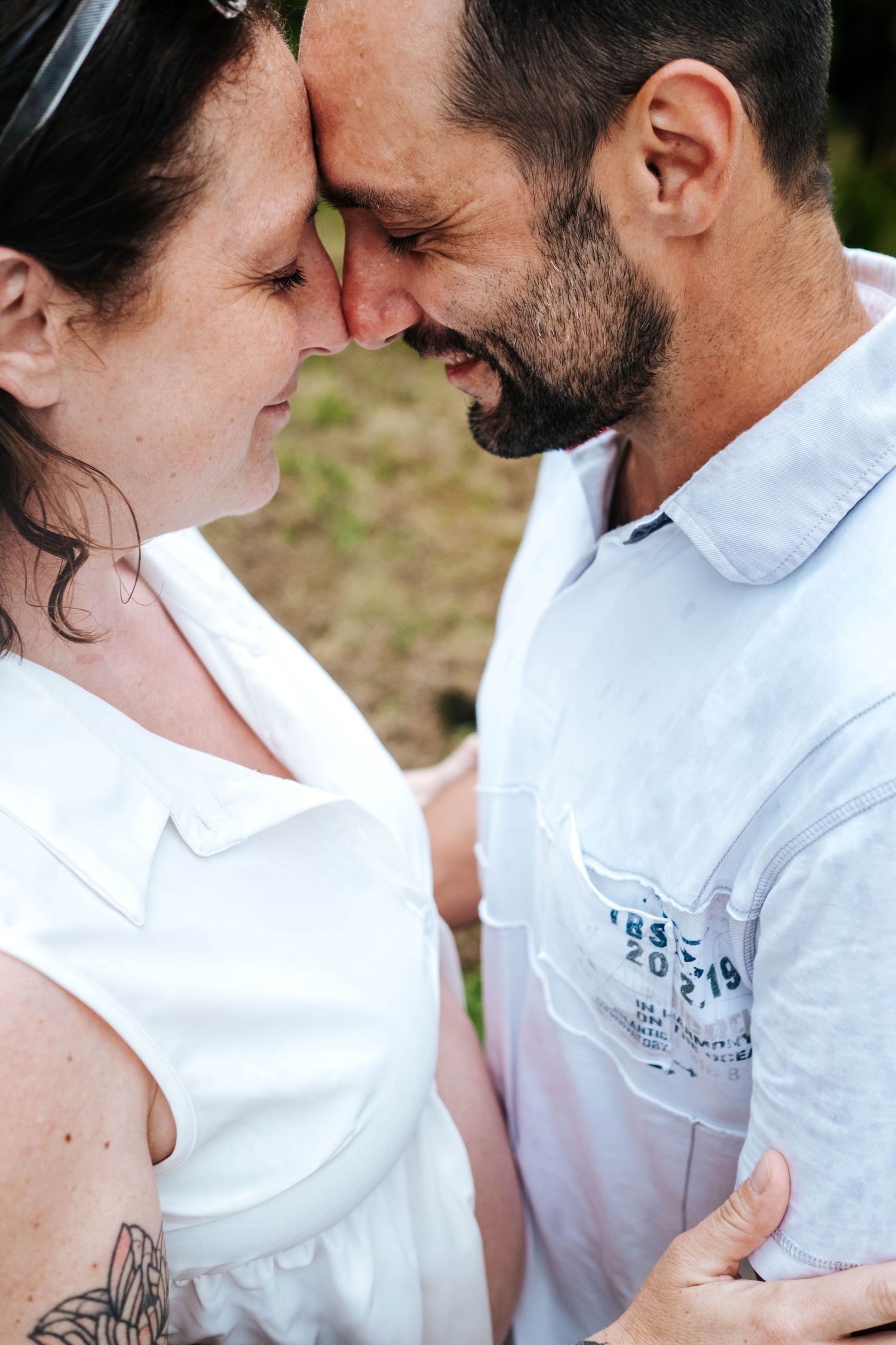 Couple sharing a tender forehead-to-forehead moment outdoors, woman in white dress with tattoo, man in light blue shirt.