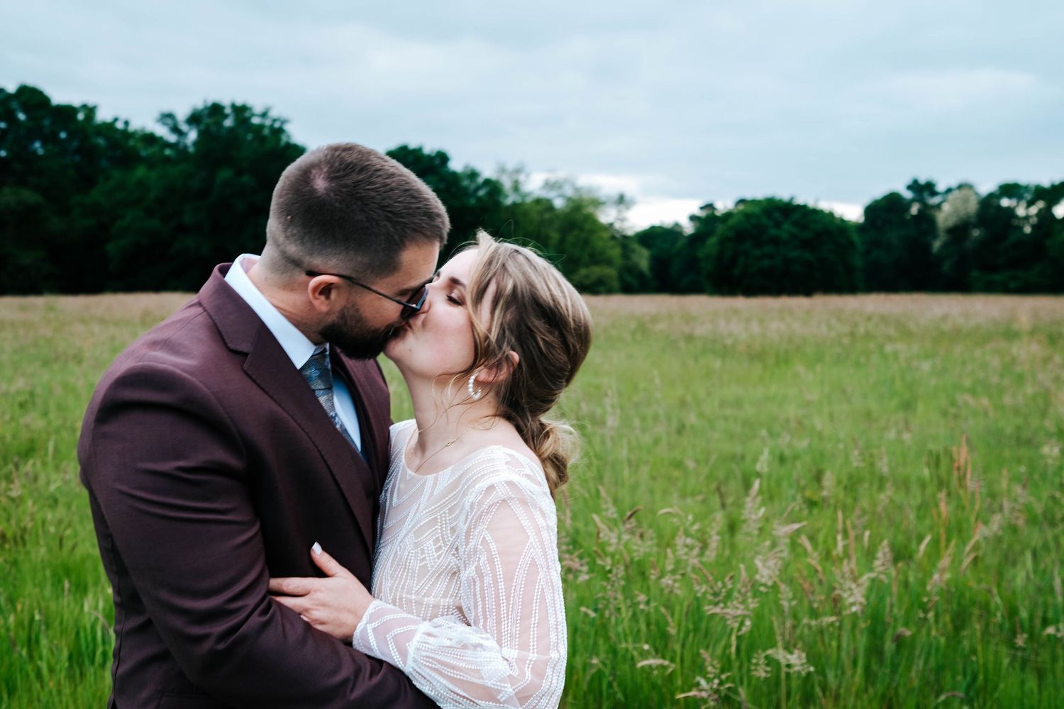 A couple shares a romantic kiss in a lush green meadow, the groom in a burgundy suit and bride in a white lace dress.