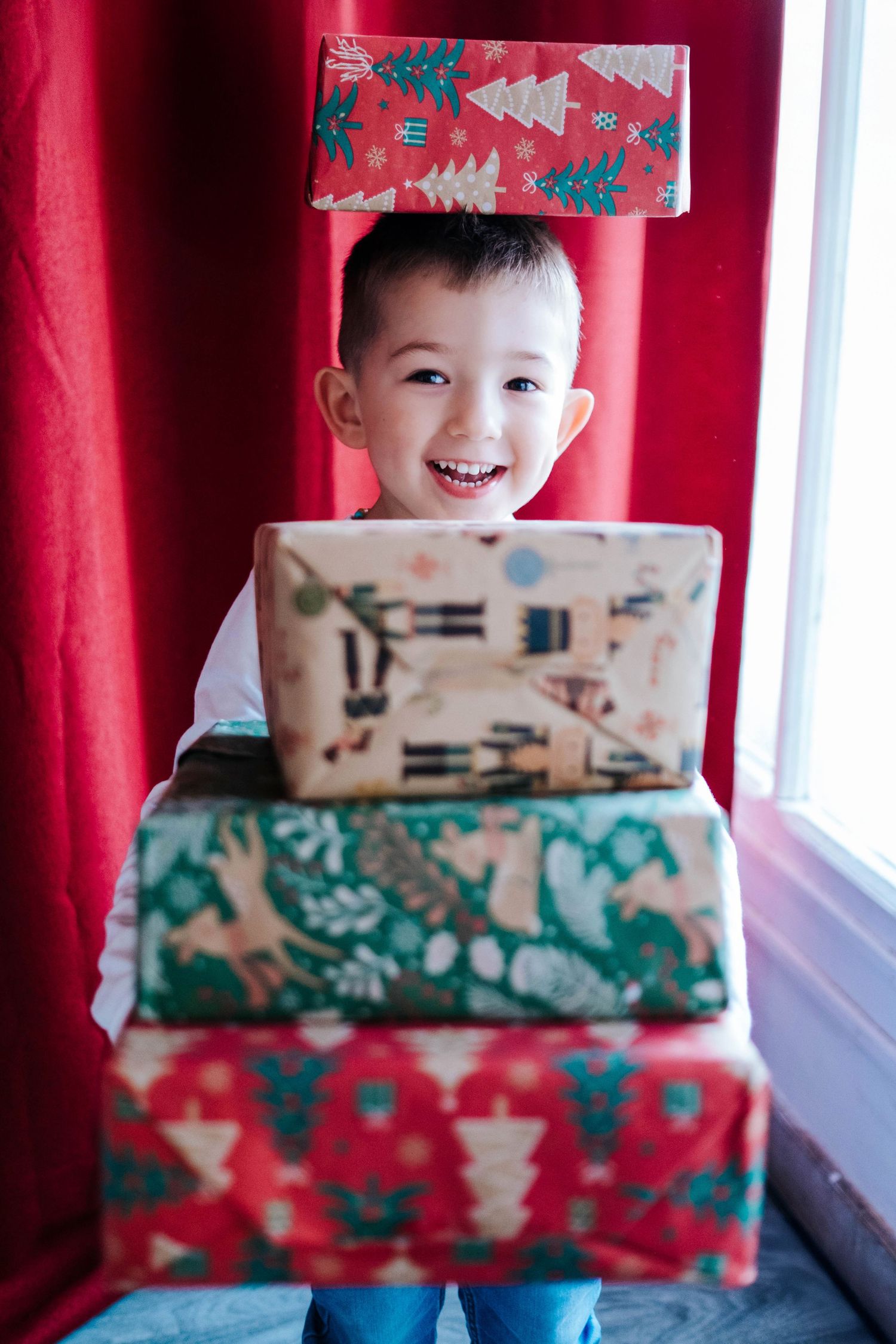 Child holds wrapped Christmas presents in front of red curtain background showing holiday excitement and joy.