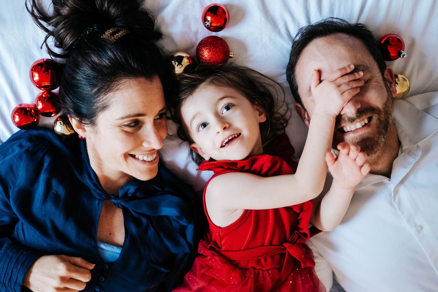 Siblings in red and blue outfits playfully pose with red Christmas ornaments on white bedding.