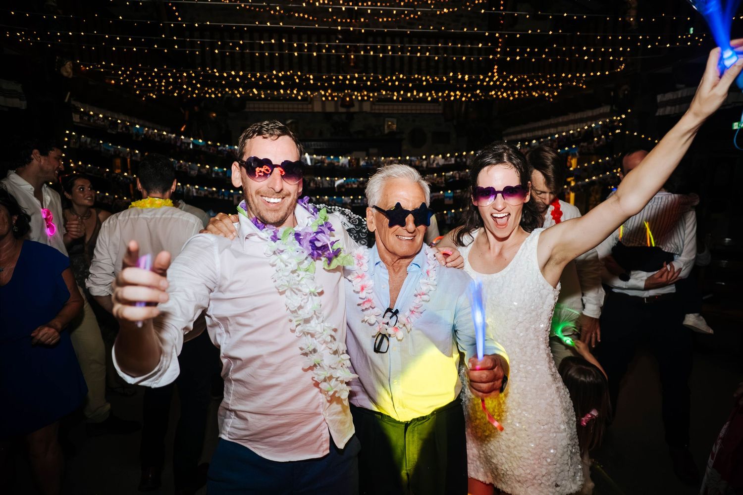 Three people in sunglasses celebrate at a festive party with string lights, leis, and glow accessories.