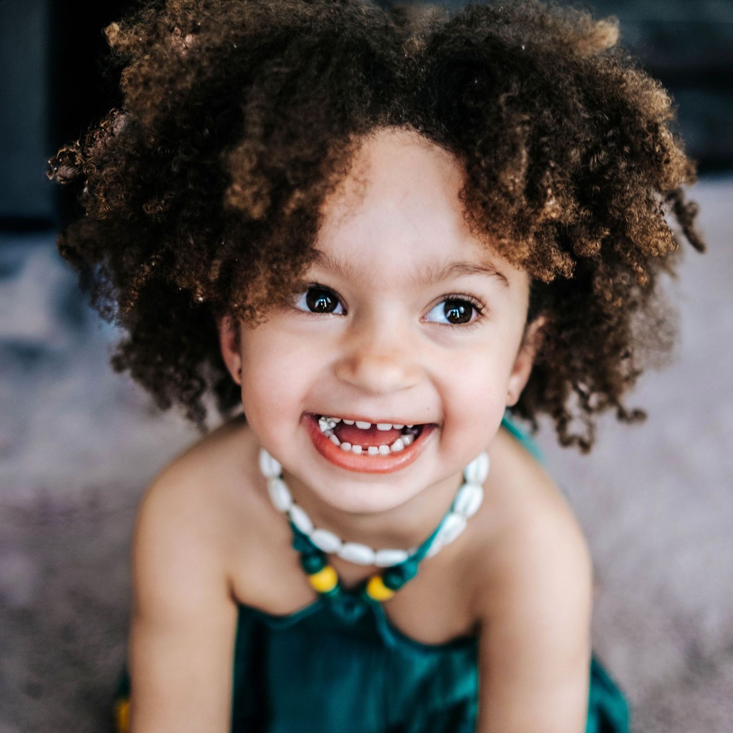 Smiling toddler girl with curly hair wearing a green dress and beaded necklace, looking up at camera.