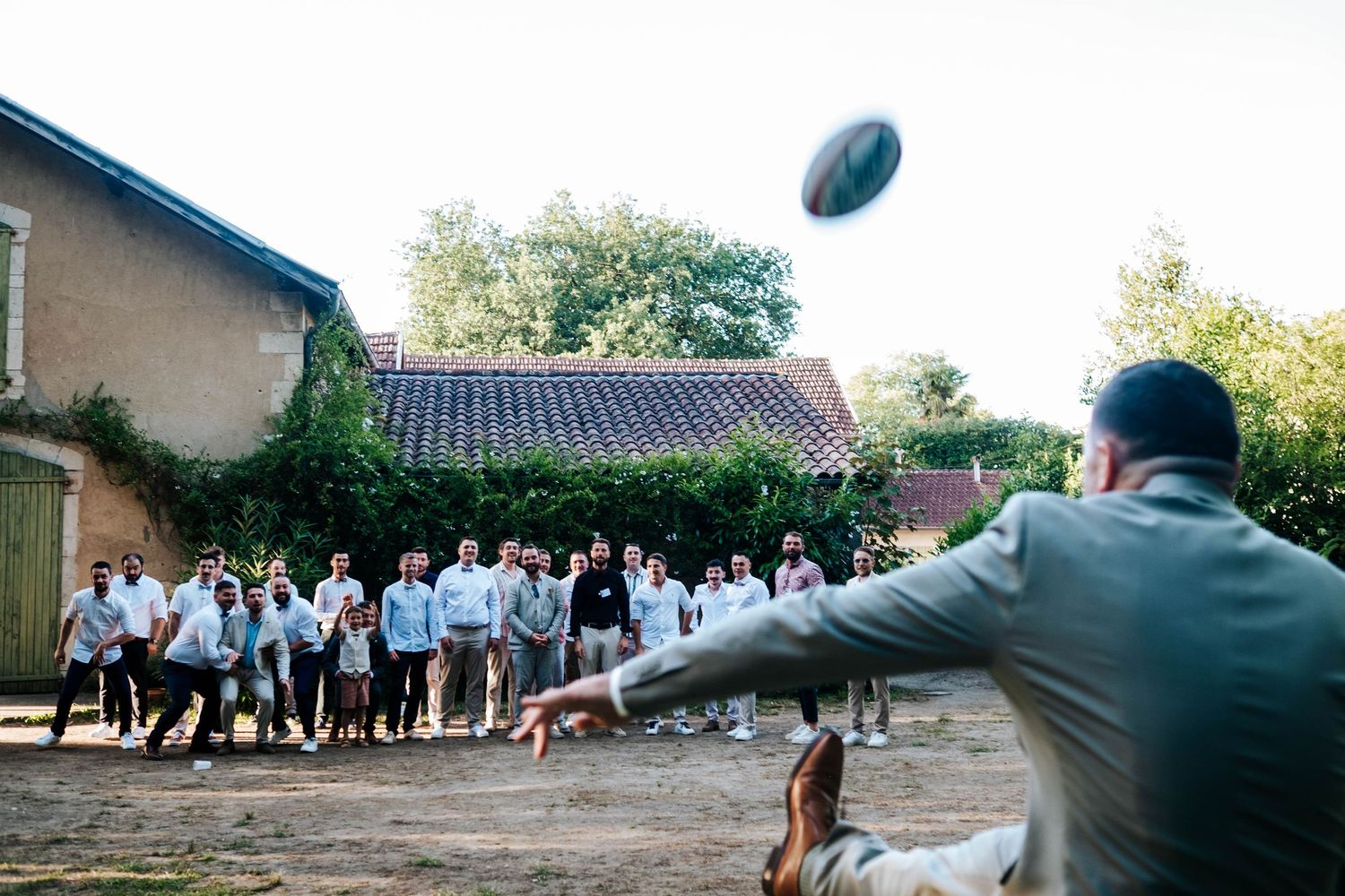 Man throwing a rugby ball at an outdoor wedding reception with guests watching near a rustic farmhouse.