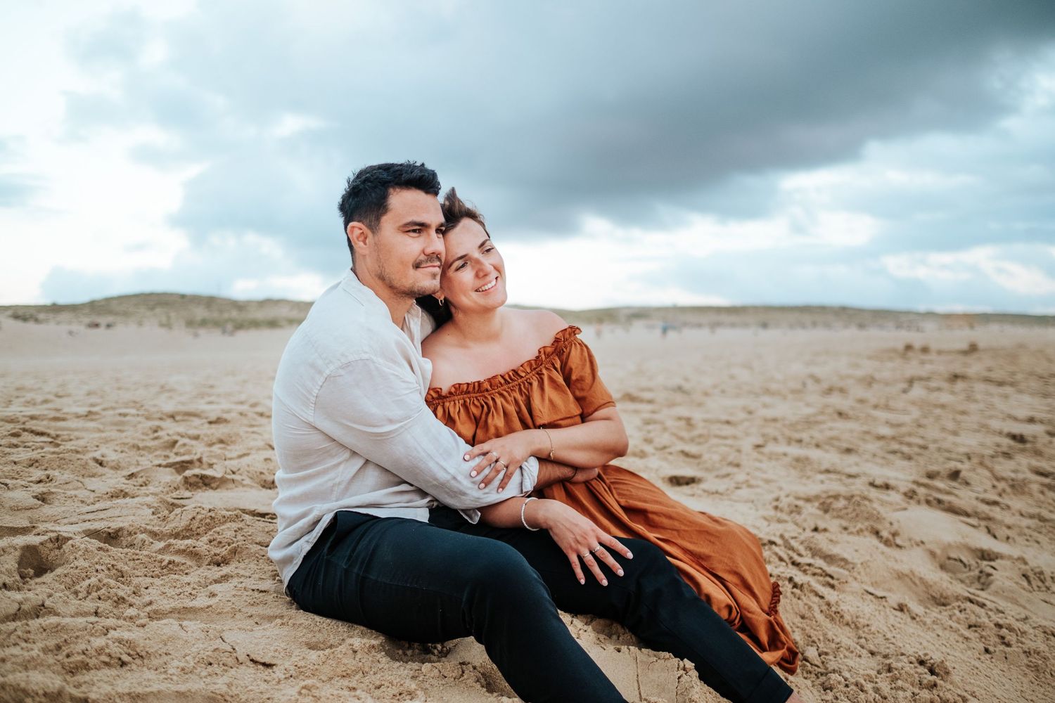 Romantic couple embraces on sandy beach under moody cloudy sky during sunset photoshoot.