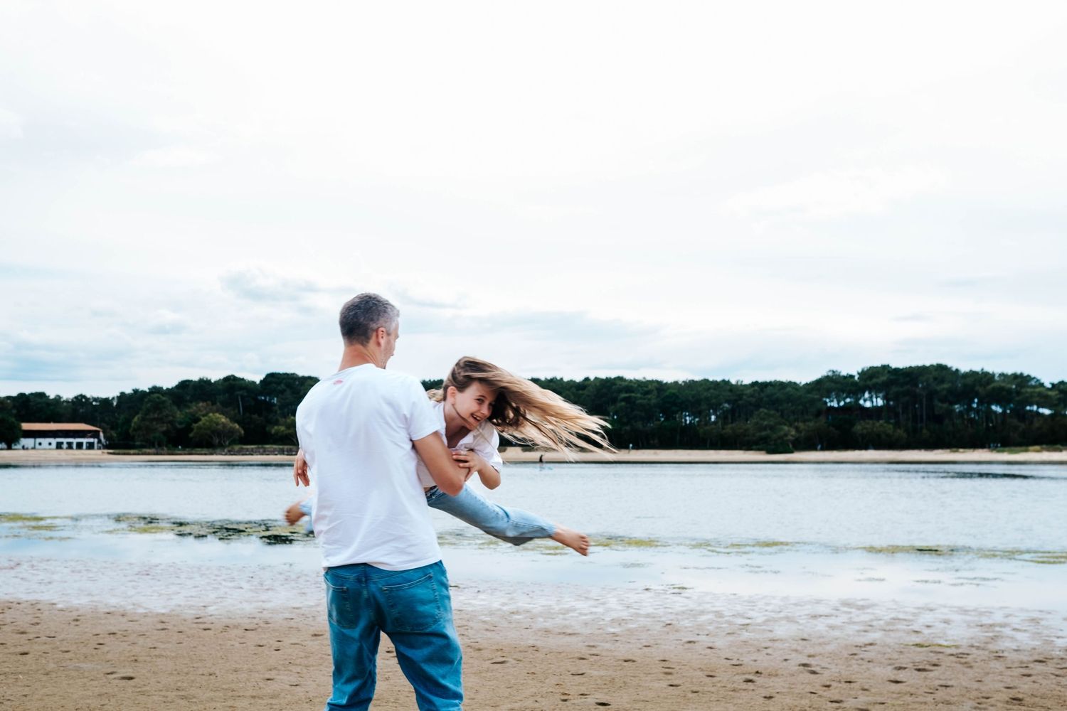 A man swings a young girl by the beach on a cloudy day, her hair flying as they enjoy a playful moment.