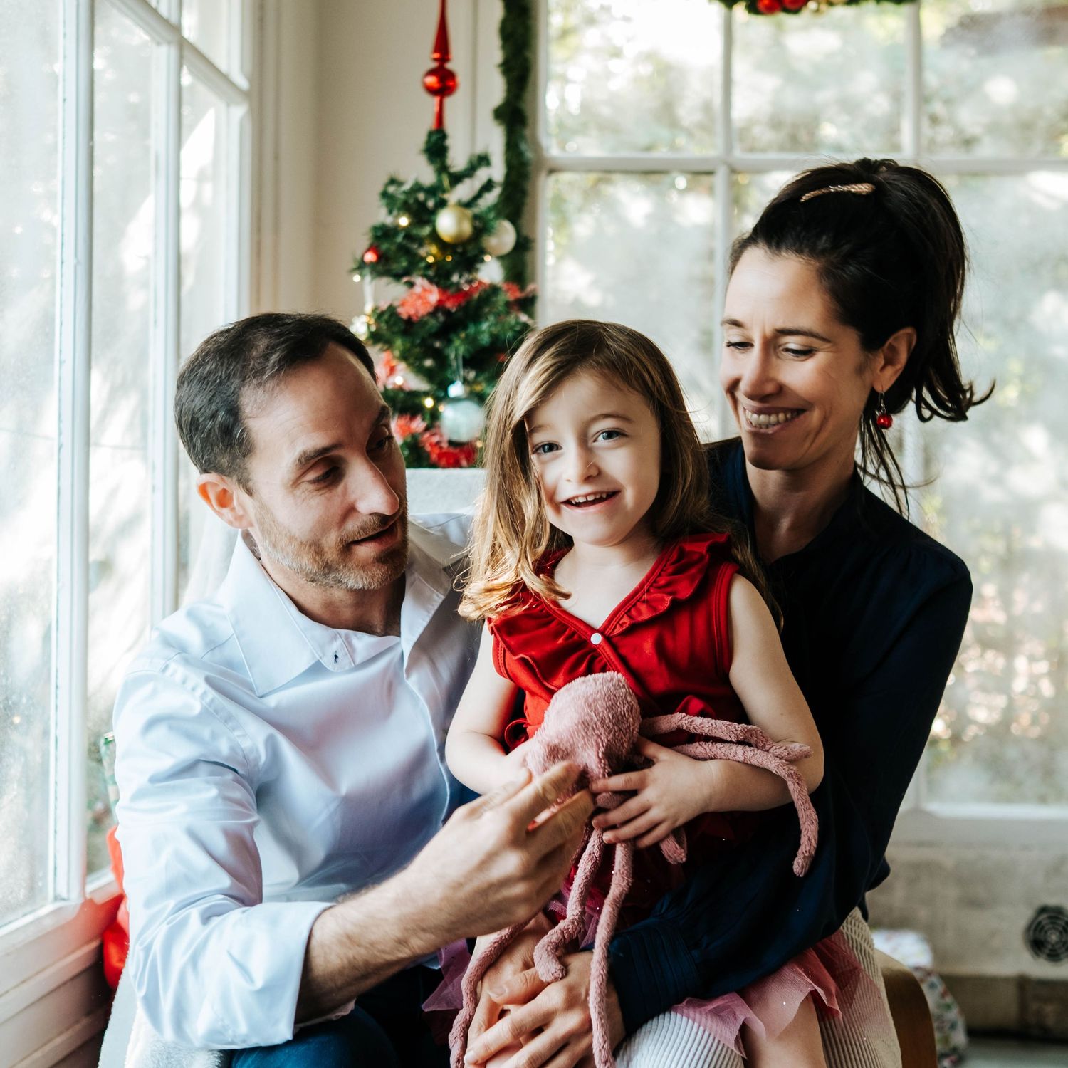 A tender family moment with a baby in red holiday attire against a bright window backdrop with Christmas decorations.