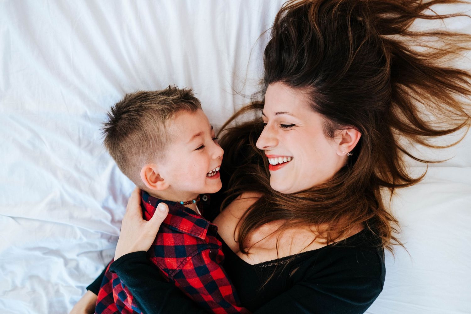 A child in a red plaid shirt and an adult with long brown hair share a joyful moment while lying on white bedding.