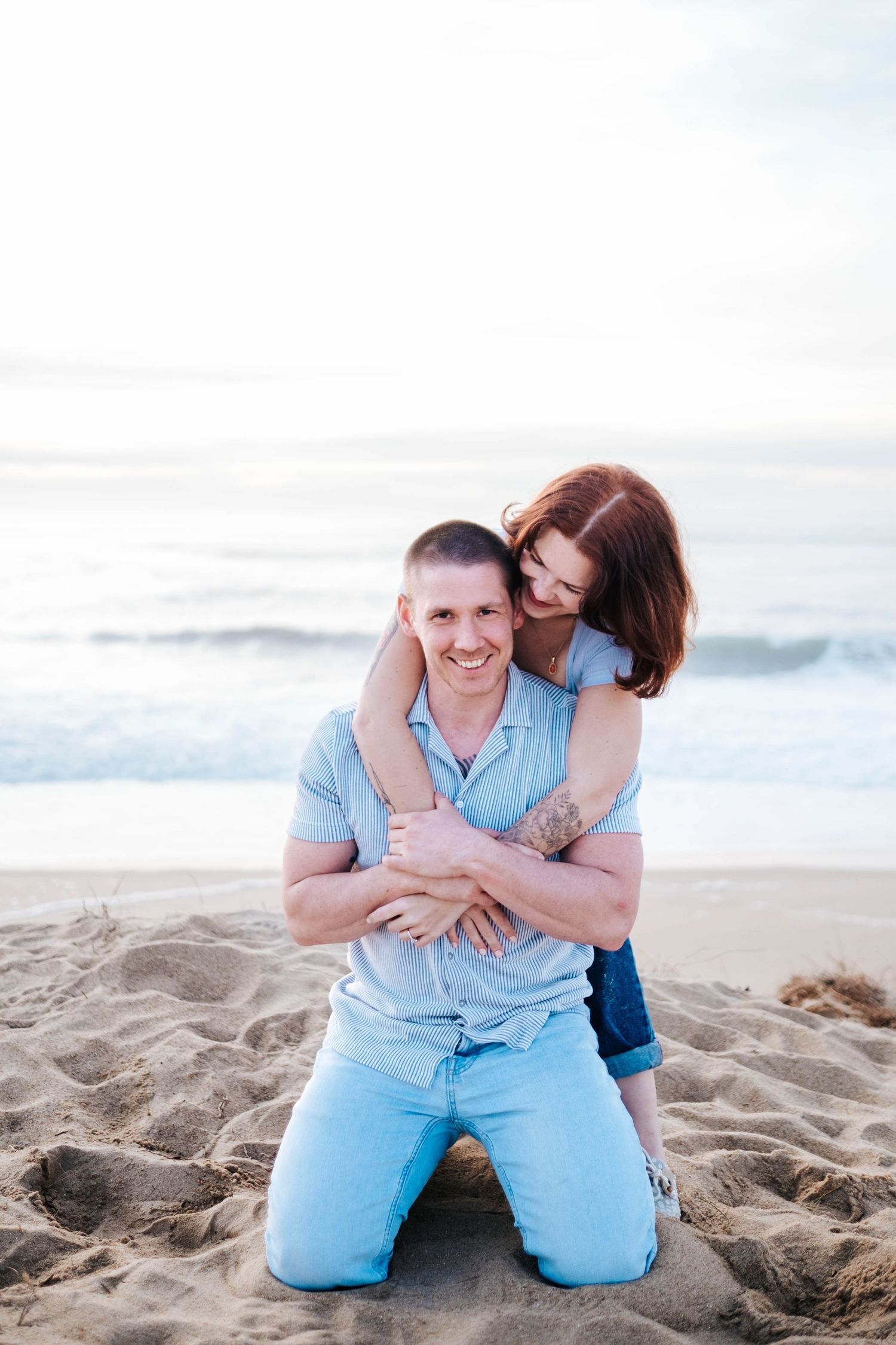 A couple embraces on a sandy beach at sunset wearing coordinating light blue outfits.