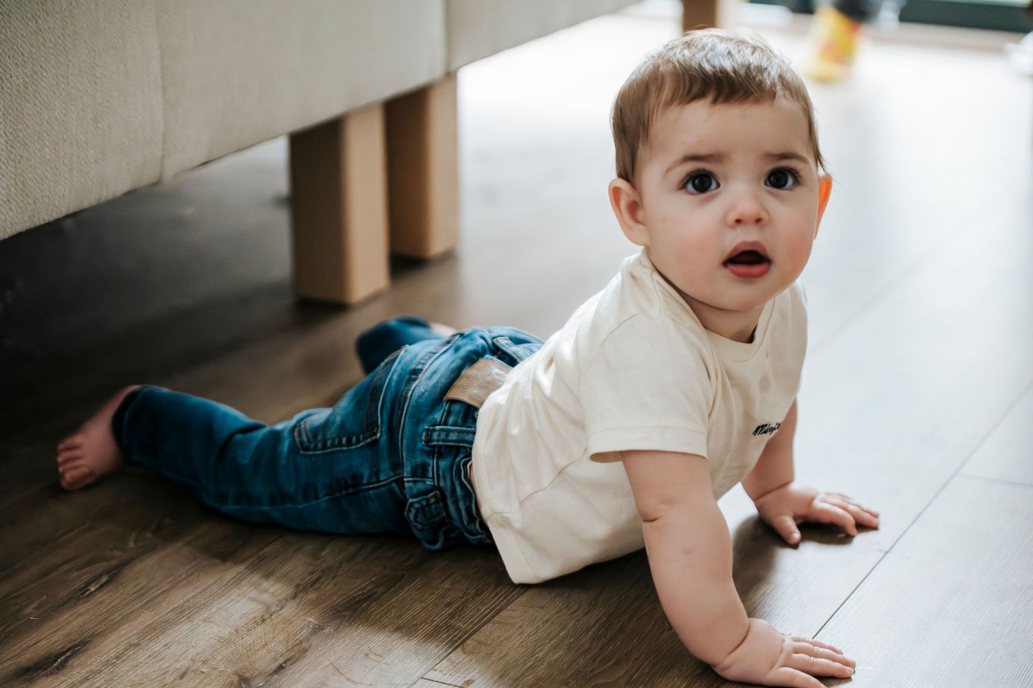 Small child in white t-shirt and jeans crawling on hardwood floor while looking back over shoulder.