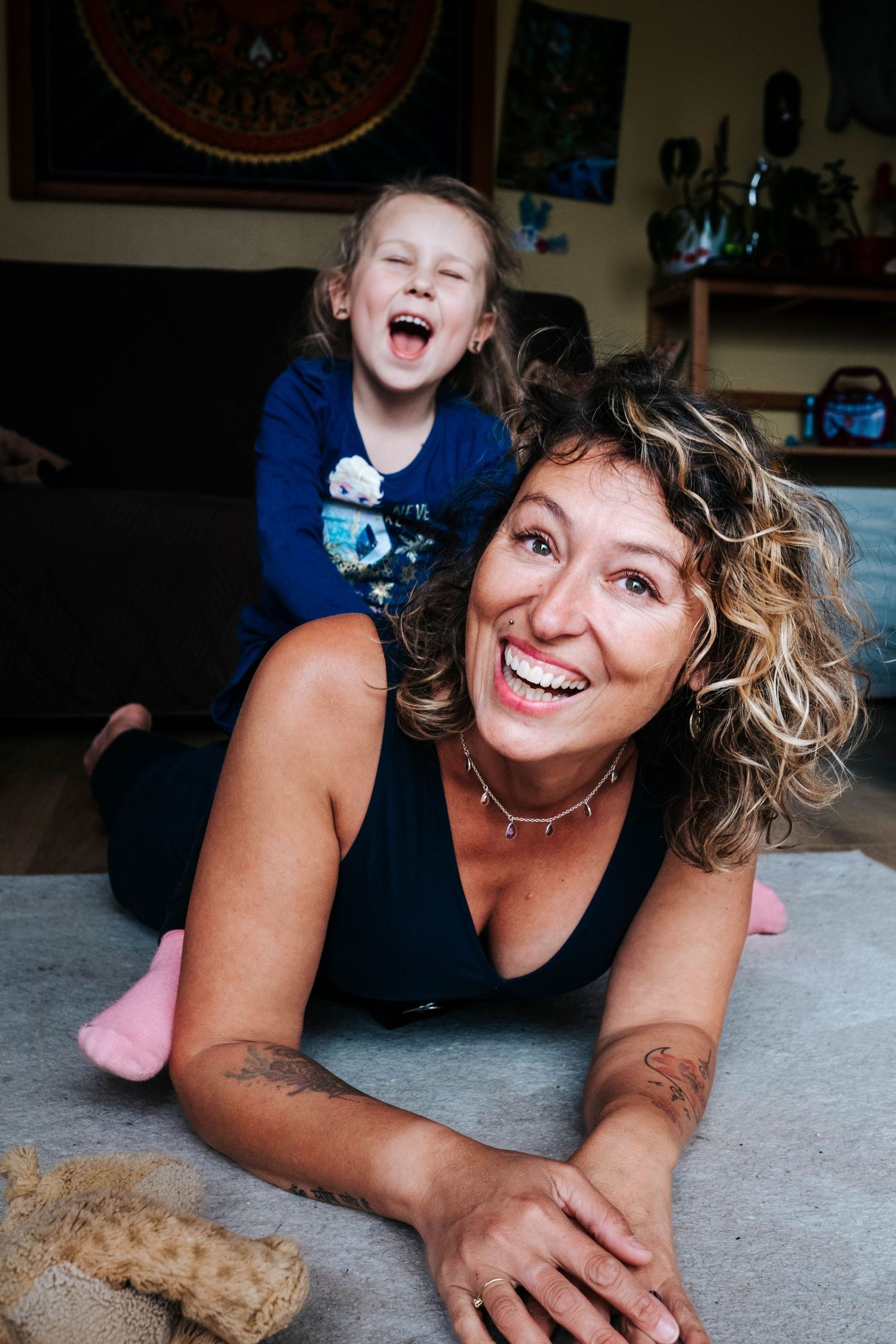 A laughing woman lying on the floor with a giggling child on her back, sharing a joyful moment at home.