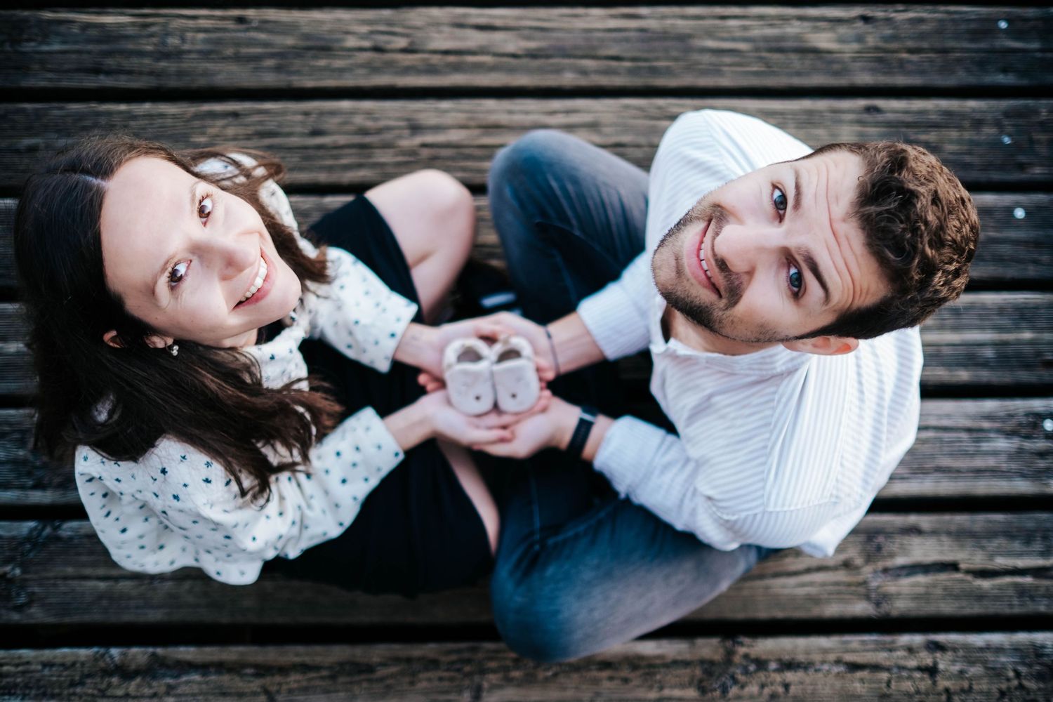 Couple sitting on wooden deck holding baby shoes, announcing pregnancy from above angle.