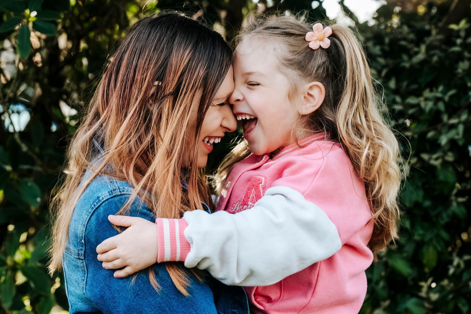 A joyful mother and young daughter laughing and hugging outdoors surrounded by green foliage.