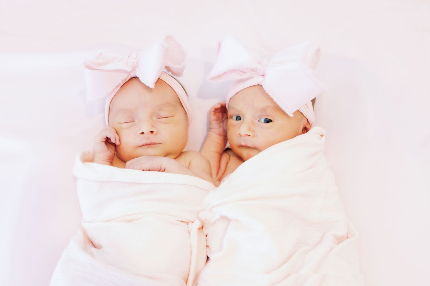 Two newborn babies swaddled in white blankets wearing pink bows lay side by side on a pale pink background.
