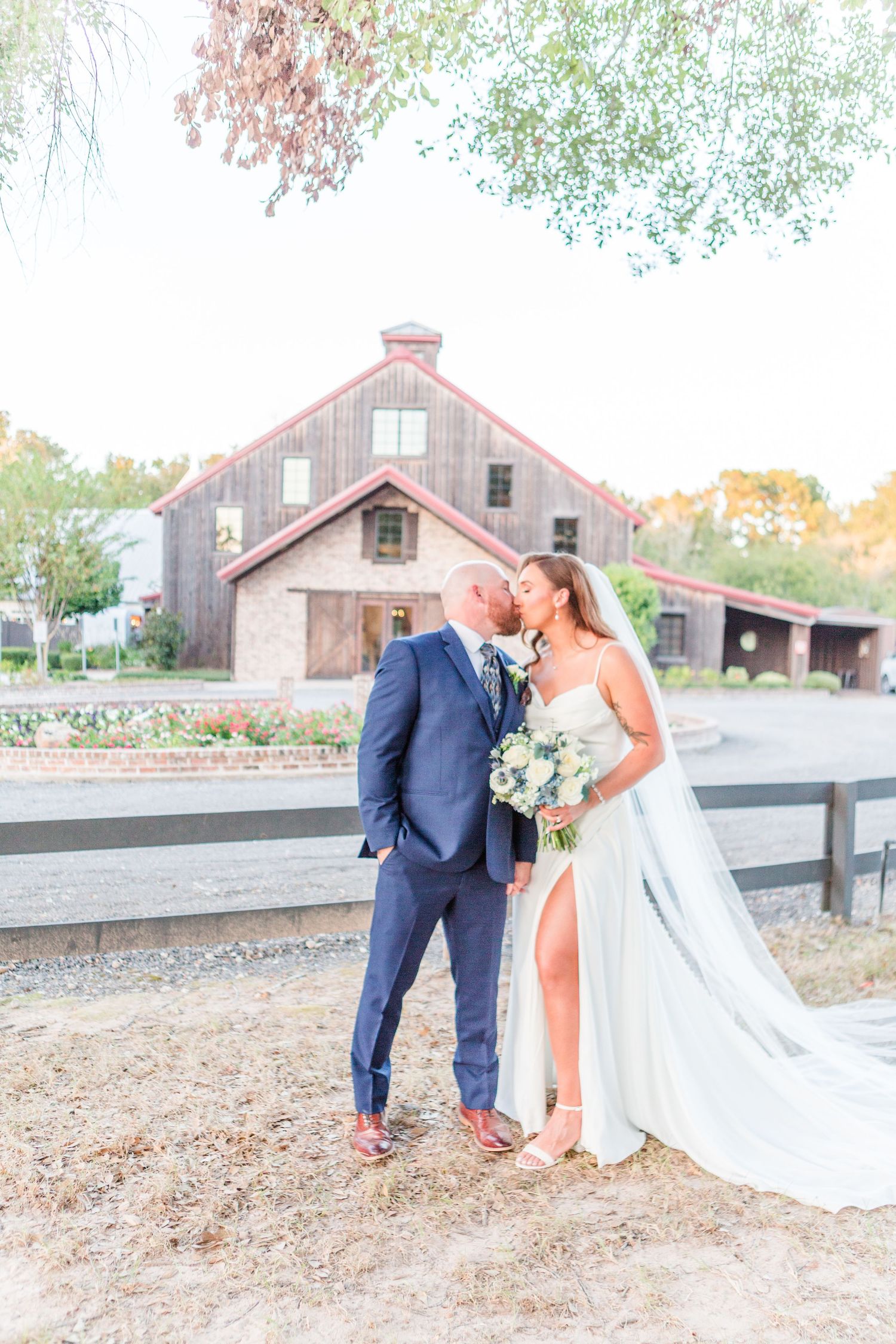 A couple in wedding attire shares romantic moments outside a rustic barn venue at sunset.
